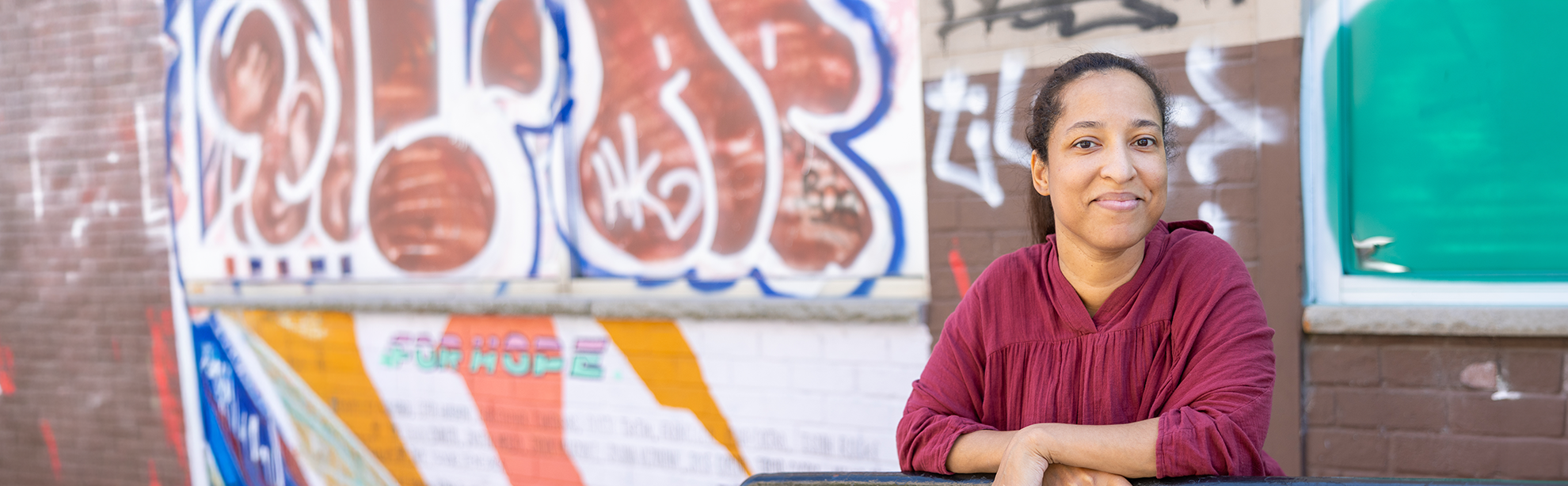 Woman leans on metal railing with hands crossed. She is smiling. Brick wall with graffiti is in the background